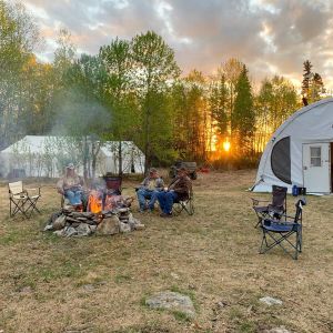 Camp setup with Quanset and large tents, and people sitting around a fire in fall at sunset