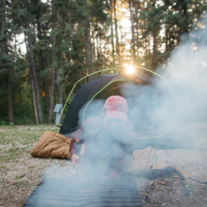 Woman camping in a tent lighting a fire in a fire pit with smoke billowing from the pit